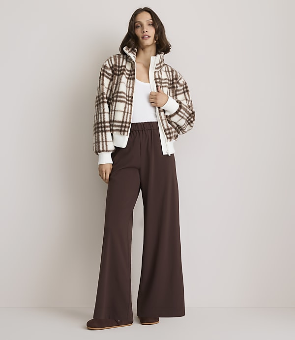 A woman in a plaid bomber jacket and wide-leg brown pants stands against a plain, light-colored background.