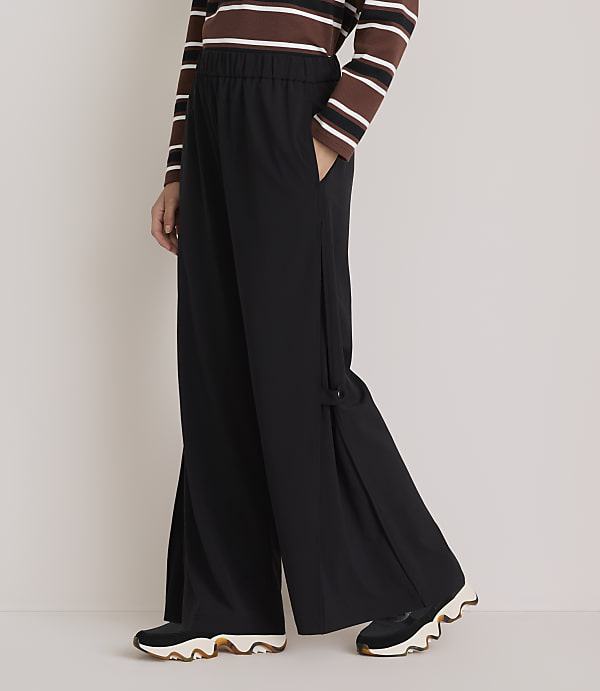 A person wearing black wide-leg pants with a striped shirt and sneakers stands against a plain gray background.
