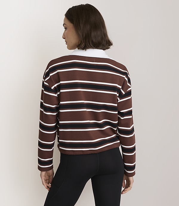 A woman in a brown, white, and black striped long-sleeved shirt and black leggings stands against a plain, light-colored background.