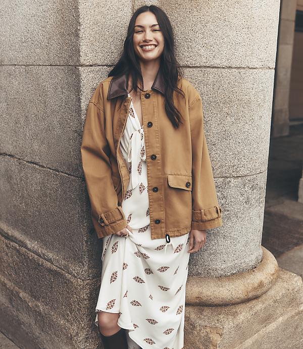 A smiling woman in a patterned dress and brown jacket leans against a stone pillar with a blurred building in the background.