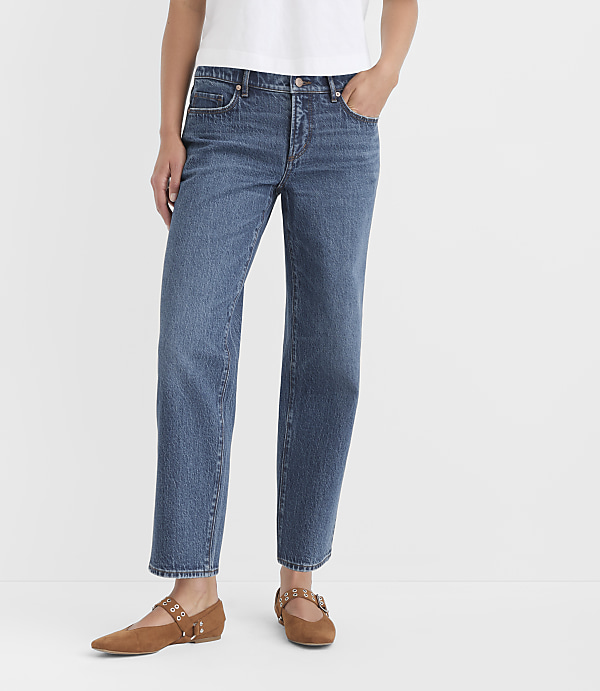 A person wearing blue jeans and brown flats stands against a plain white background.