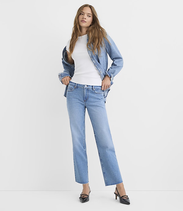 A woman in a white top, denim jacket, and jeans stands against a plain white background.