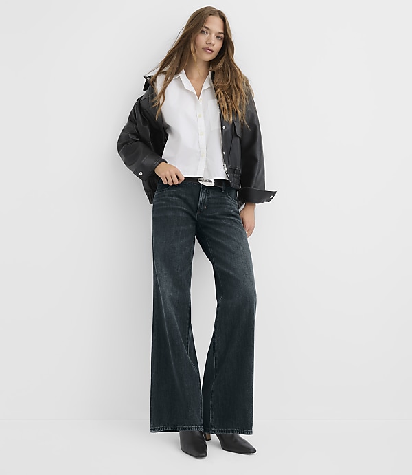 A woman in a white shirt, black jacket, and wide-leg jeans stands against a plain white background.