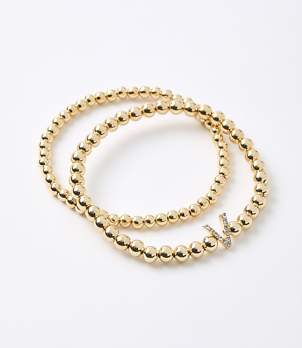 Two gold beaded bracelets, one with a diamond-encrusted "V" charm, rest on a plain white background.