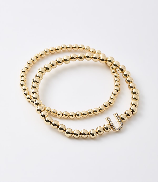 Two gold beaded bracelets, one with a diamond-encrusted "U" charm, lie on a plain white background.