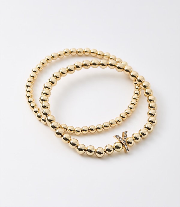 Two gold beaded bracelets, one with a diamond-studded "X" charm, are arranged on a plain white background.
