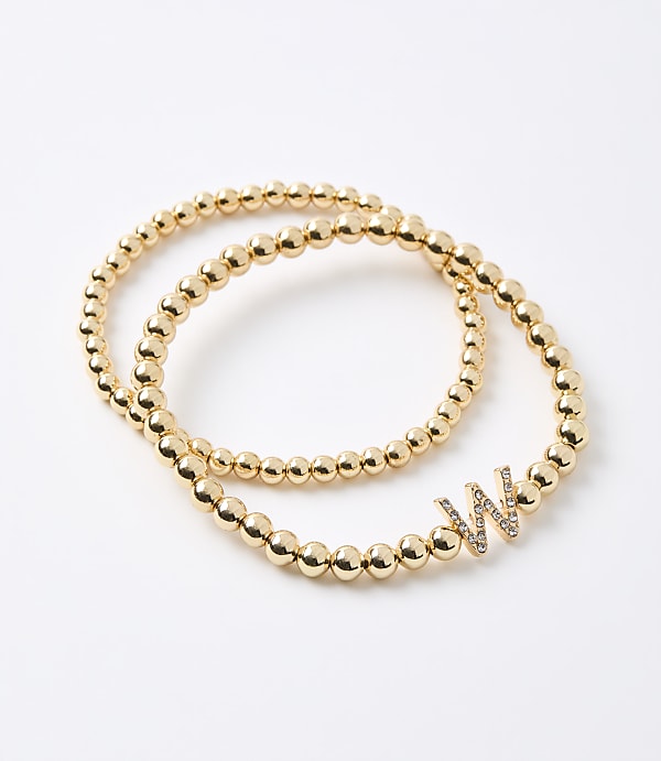 Two gold beaded bracelets, one with a diamond-encrusted "W" charm, rest on a plain white background.