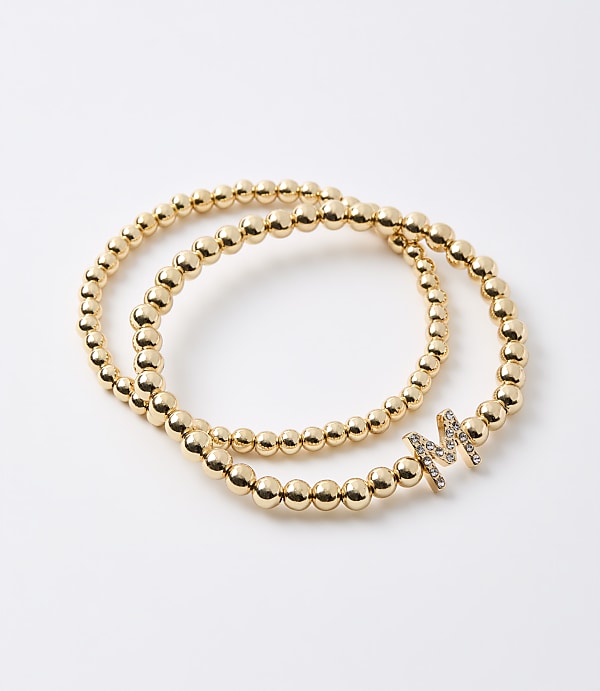 Two gold beaded bracelets, one with a diamond-encrusted letter "M" charm, rest on a plain white background.
