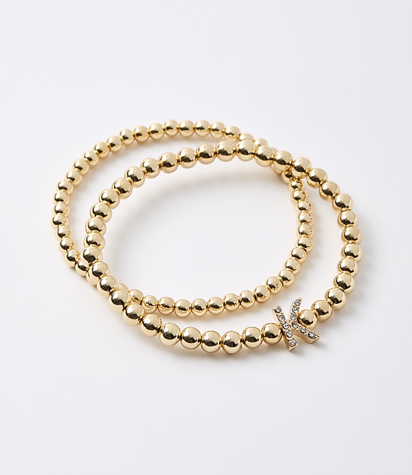 Two gold beaded bracelets, one wrapped around the other, lie on a plain white background, with one bracelet featuring a small, diamond-encrusted letter "K" charm.