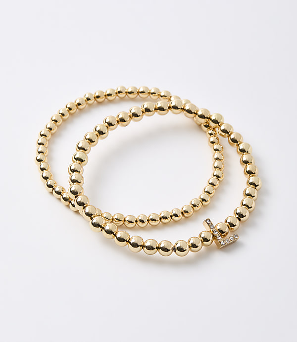 Two gold beaded bracelets, one with a diamond-encrusted initial "L" charm, rest on a plain white background.