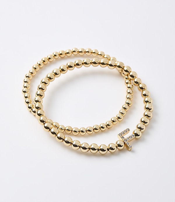 A gold beaded bracelet with a diamond-encrusted letter "F" charm rests on a plain white background.