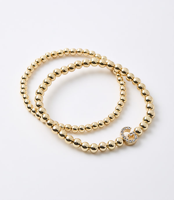 Two gold beaded bracelets, one with a diamond-encrusted infinity charm, lie on a plain white background.