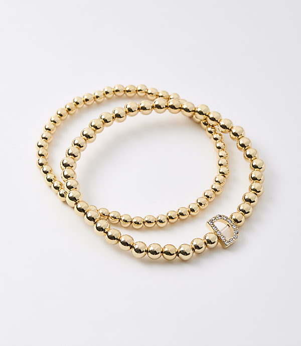 Two gold beaded bracelets, one with a diamond-encrusted "D" charm, lie on a plain white background.