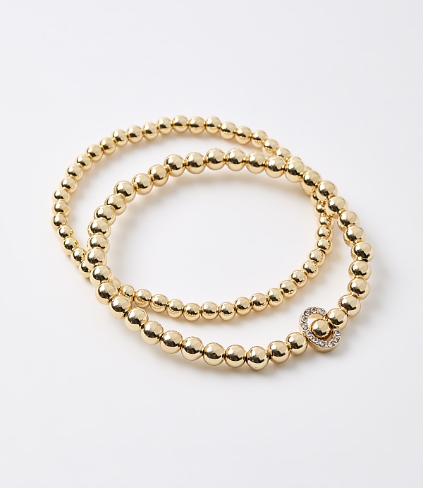 A gold beaded bracelet with a clover-shaped charm rests on a plain white background.