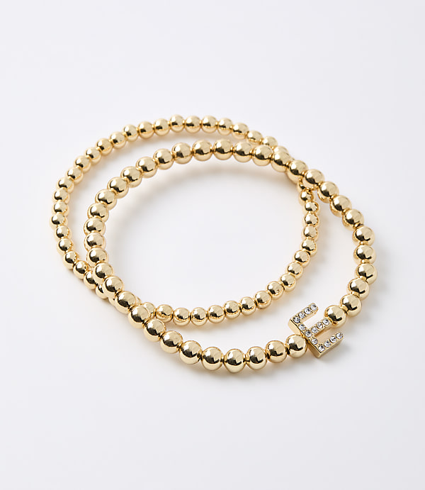 Two gold beaded bracelets, one with a diamond-encrusted letter "E" charm, lie on a plain white background.