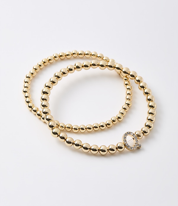 Two gold beaded bracelets, one with a diamond-encrusted "Q" charm, rest on a plain white background.