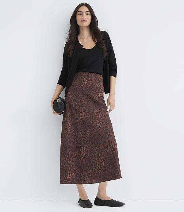 A woman in a leopard print skirt and black top stands against a plain white background, holding a small black purse.
