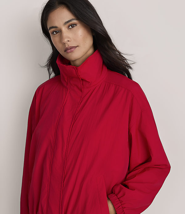 A woman in a red jacket stands against a plain, light-colored background.