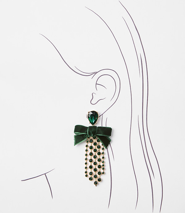 A green velvet bow earring with a teardrop gem and dangling rhinestones hangs from a stylized ear against a plain white background.