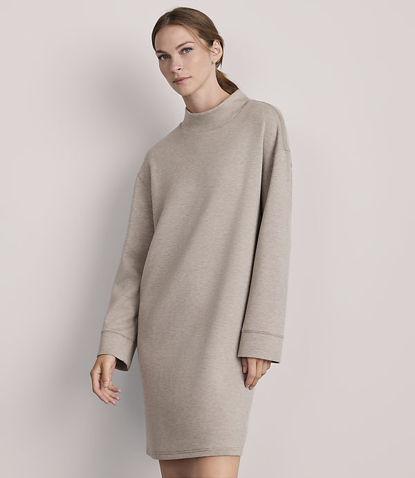 A woman in a beige sweatshirt dress stands against a plain, light gray background.