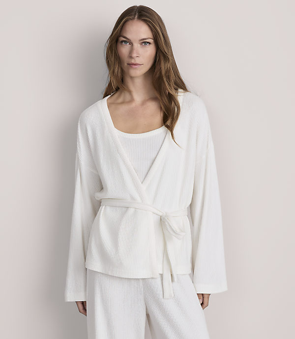 A woman in a white ribbed robe and matching pants stands against a plain, light-colored background.