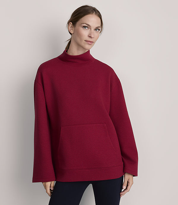 A woman in a red waffle-knit turtleneck sweater with a front pocket stands against a plain gray background.
