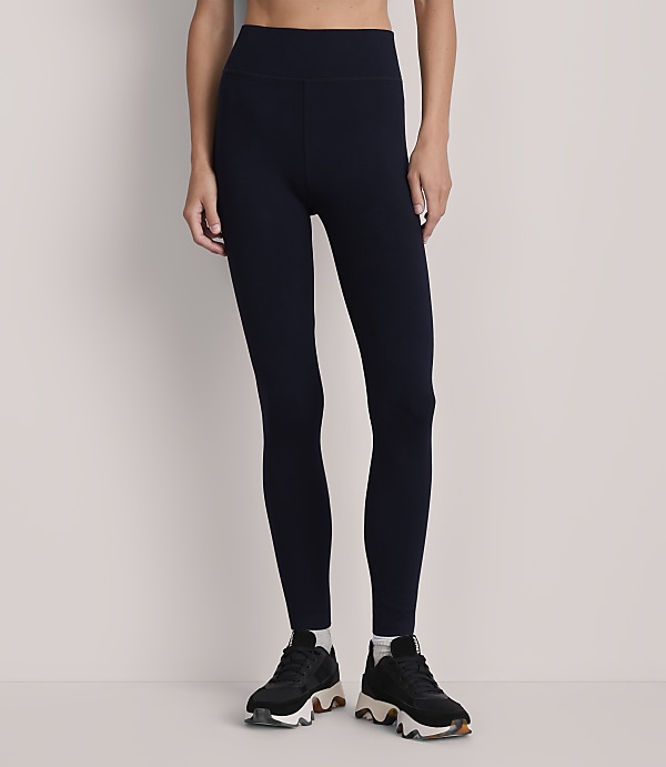 A person stands in dark blue leggings and black sneakers against a plain, light gray background.
