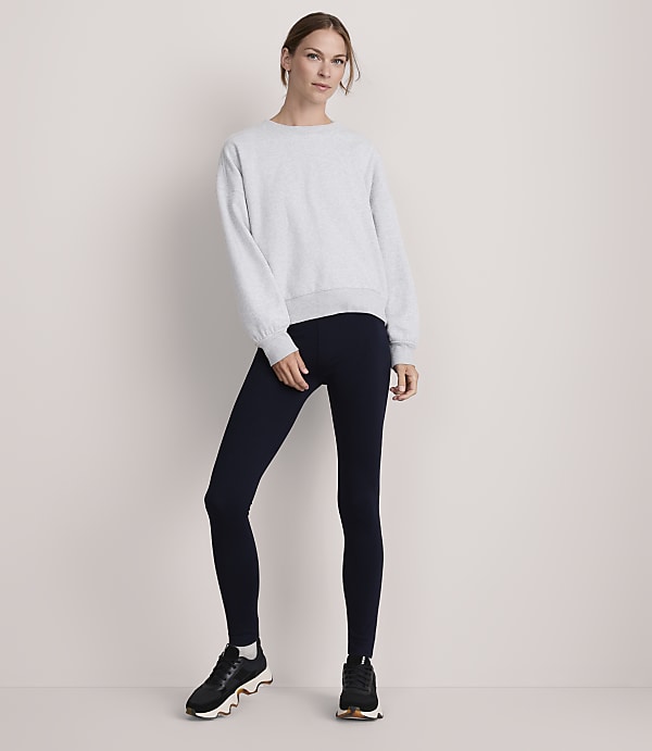 A woman in a light gray sweatshirt and dark blue leggings stands against a plain, light-colored background.