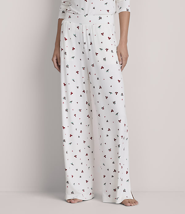 A person stands in white, wide-leg pajama pants adorned with small red and green floral patterns, against a plain, light gray background.