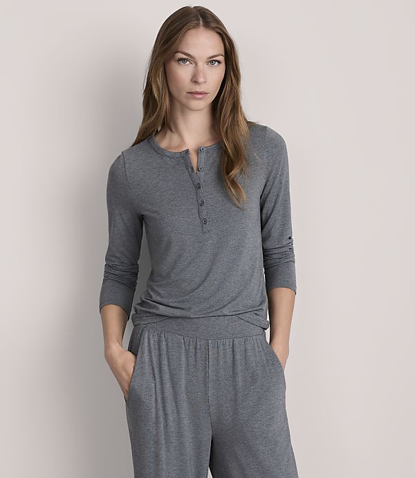 A woman in a grey henley shirt and matching pants stands against a plain, light grey background.