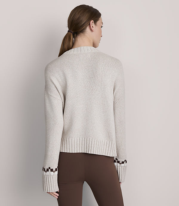 A woman in a light-colored sweater and brown leggings stands against a plain gray background.