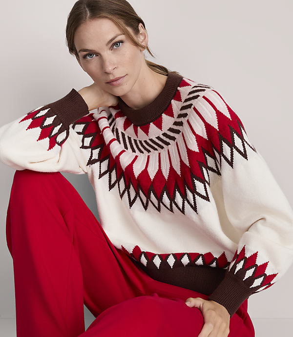 A woman in a cream-colored sweater with a red and brown geometric pattern poses against a plain background.