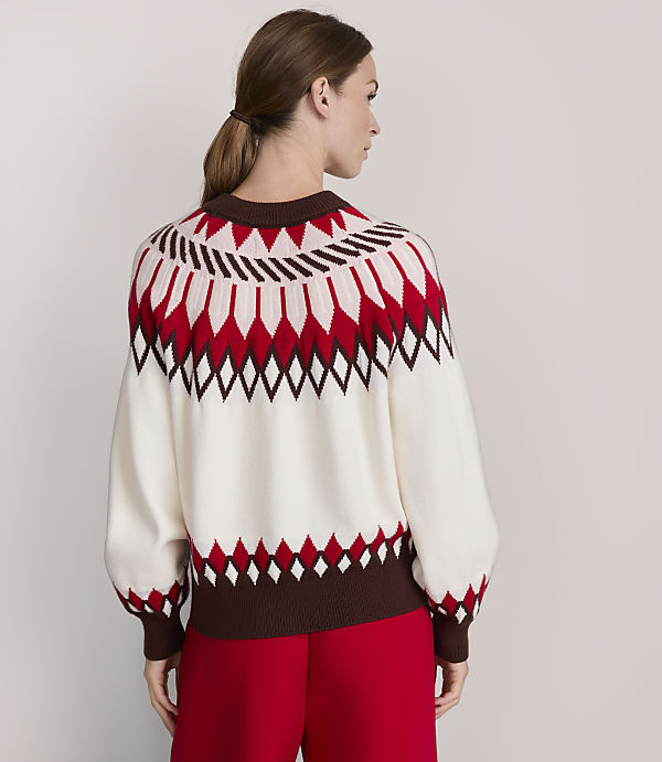 A woman in a cream, red, and brown patterned sweater stands against a plain, light gray background.