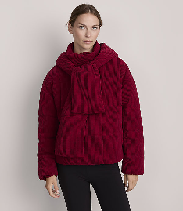 A woman in a red puffer jacket and scarf stands against a plain gray background.