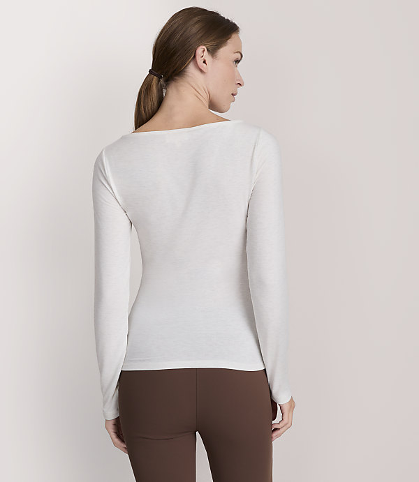 A woman in a white long-sleeved shirt and brown leggings stands against a plain white background.