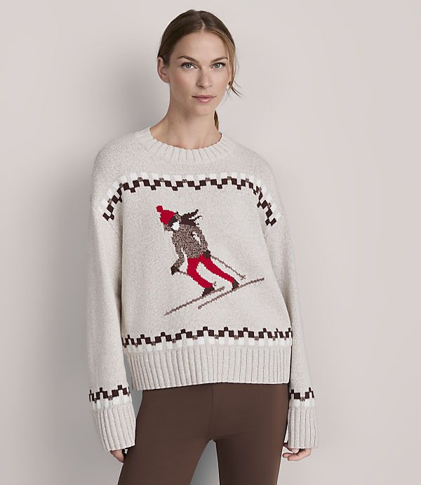 A woman in a cream-colored sweater with a skiing graphic stands against a plain, light gray background.