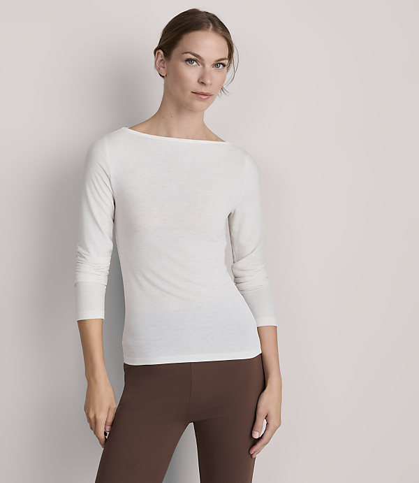 A woman in a white boatneck shirt and brown leggings stands against a plain, light gray background.