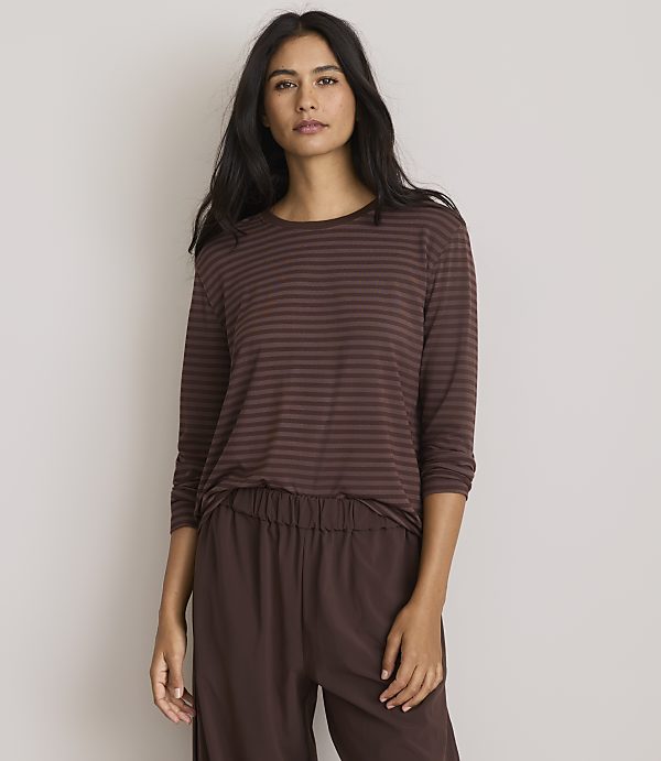 A woman with long dark hair stands in the foreground wearing a brown striped long-sleeved shirt and brown pants, against a plain, light gray background.