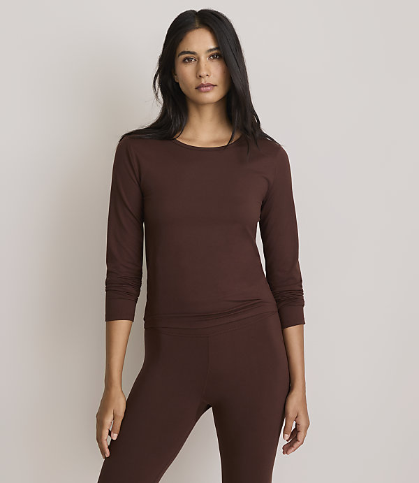 A woman in a brown long-sleeved shirt and leggings stands against a plain, light gray background.