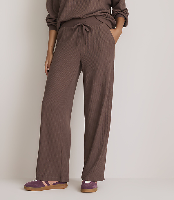 A person stands in brown waffle-knit pants and purple sneakers against a plain, light background.