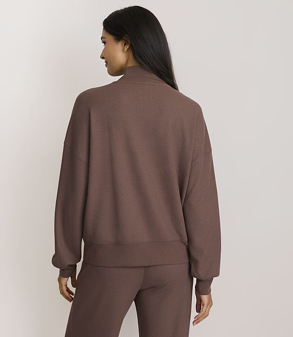 A woman in a brown ribbed sweatshirt and pants stands against a plain, light brown background.