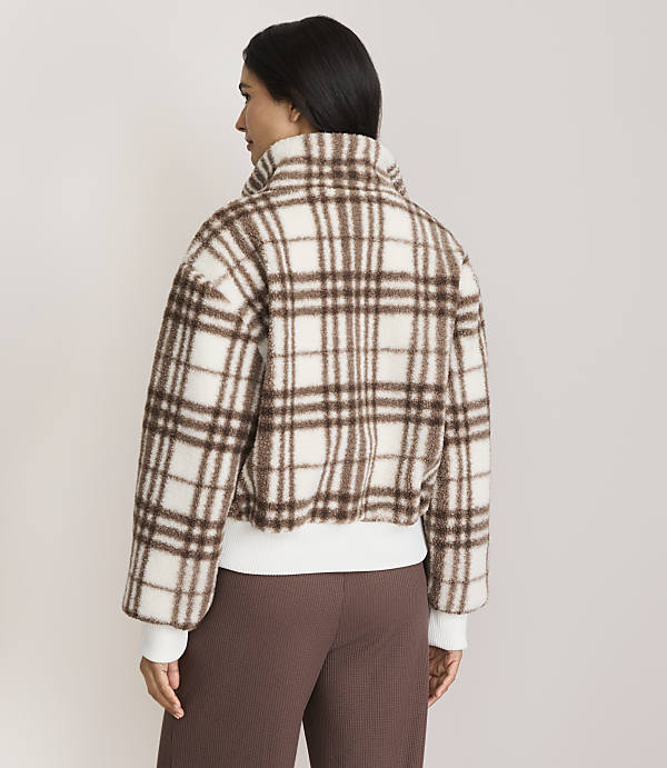 A woman in a brown and white plaid jacket stands against a plain, light-colored background.