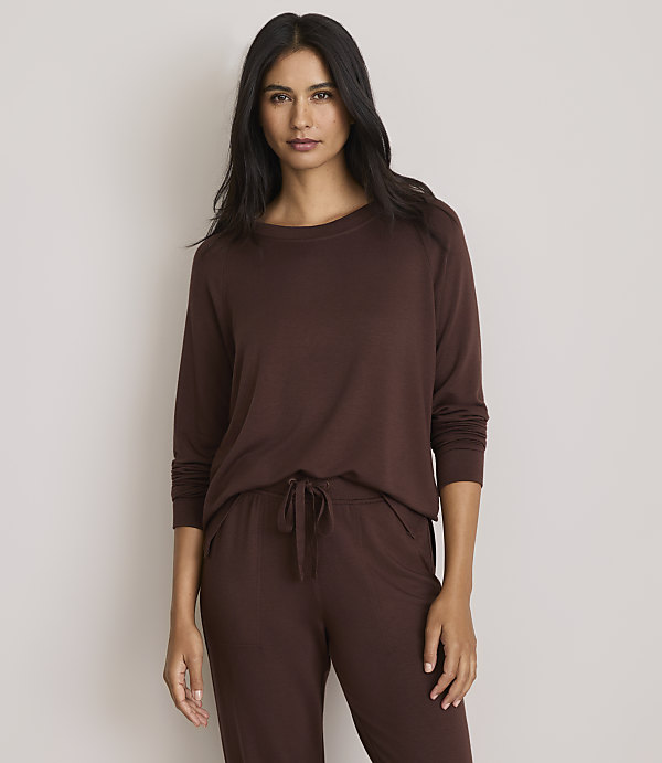 A woman in a brown long-sleeved shirt and pants stands against a plain, light gray background.