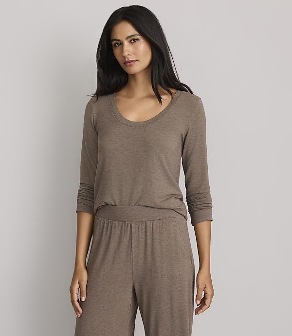 A woman in a brown long-sleeved shirt and matching pants stands against a plain gray background.