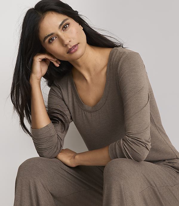 A woman in a brown long-sleeved shirt and pants rests her head on her hand against a plain gray background.
