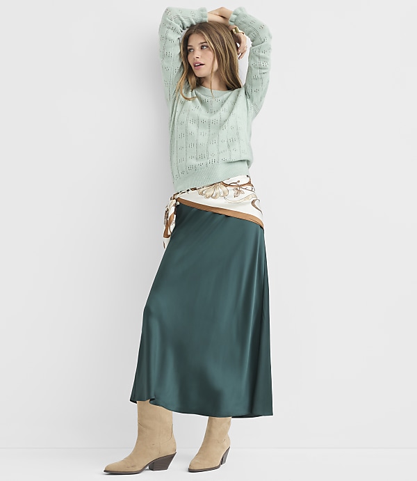 A woman in a light green sweater and a dark green satin skirt stands against a plain white background.