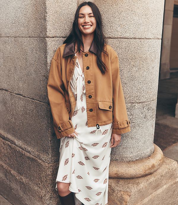 A smiling woman in a patterned dress and brown jacket leans against a stone pillar with a blurred building in the background.