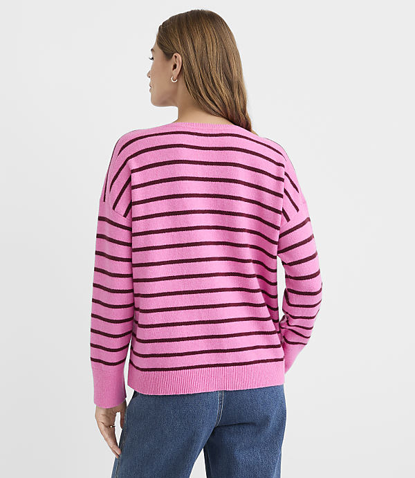 A woman in a pink and burgundy striped sweater and blue jeans stands against a plain white background.