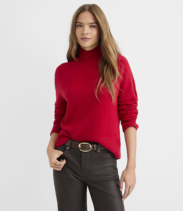 A woman in a red turtleneck sweater and dark pants stands against a plain white background.