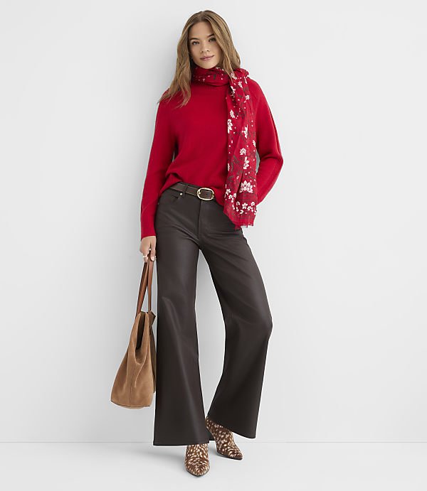 A woman in a red turtleneck and dark flared pants stands against a plain white background, holding a brown tote bag and wearing a patterned scarf and animal-print boots.